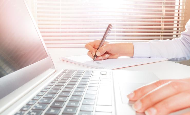 A person using a laptop and taking notes. Image by Sergey Novikov via Shutterstock.