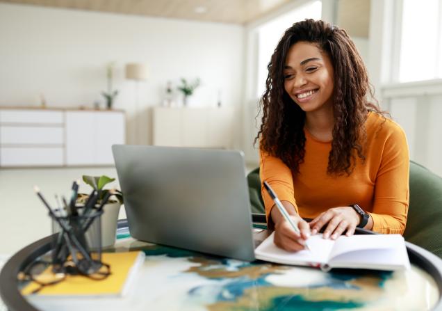 Woman sitting at desk working on laptop taking notes. Image by Prostock-studio via Shutterstock.