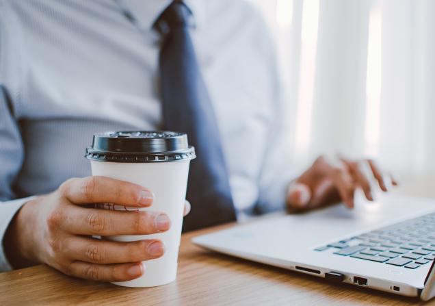 A man in a suit and tie looking at something on a laptop. He is holding a paper cup in his right hand. Image by Lisa Fotios via Pexels.