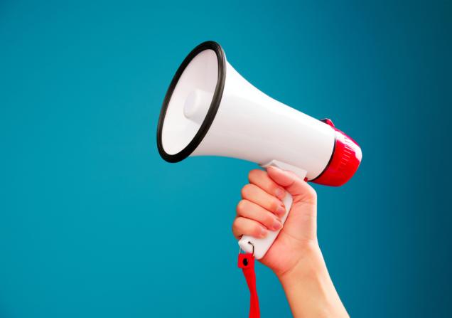 A picture of a hand holding up a megaphone against a turquoise background. Image by Sergey Mironov via Shutterstock