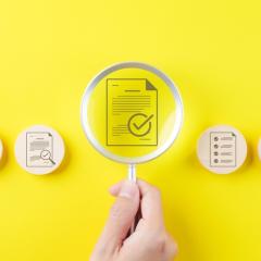 Image by SuPatMaN via Shutterstock. In the centre of the image, a hand is holding a magnifying glass over an icon of a document with a tick mark on it. On either side are more icons with documents and tick marks on wooden circles. The background of the image is bright yellow. Image by SuPatMaN via Shutterstock.