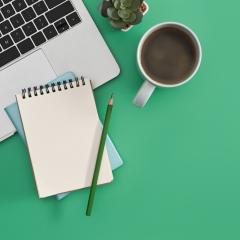 Image by Prathankarnpap via Shutterstock. A laptop, spiral-bound notepad, pencil, cup of coffee and a small plant are seen from above. The background is green. Image by Prathankarnpap via Shutterstock.