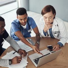 Image by PeopleImages.com - Yuri A via Shutterstock. Three people sitting round a table looking at a laptop. Two of them have stethoscopes around their neck. Image by PeopleImages.com - Yuri A via Shutterstock.